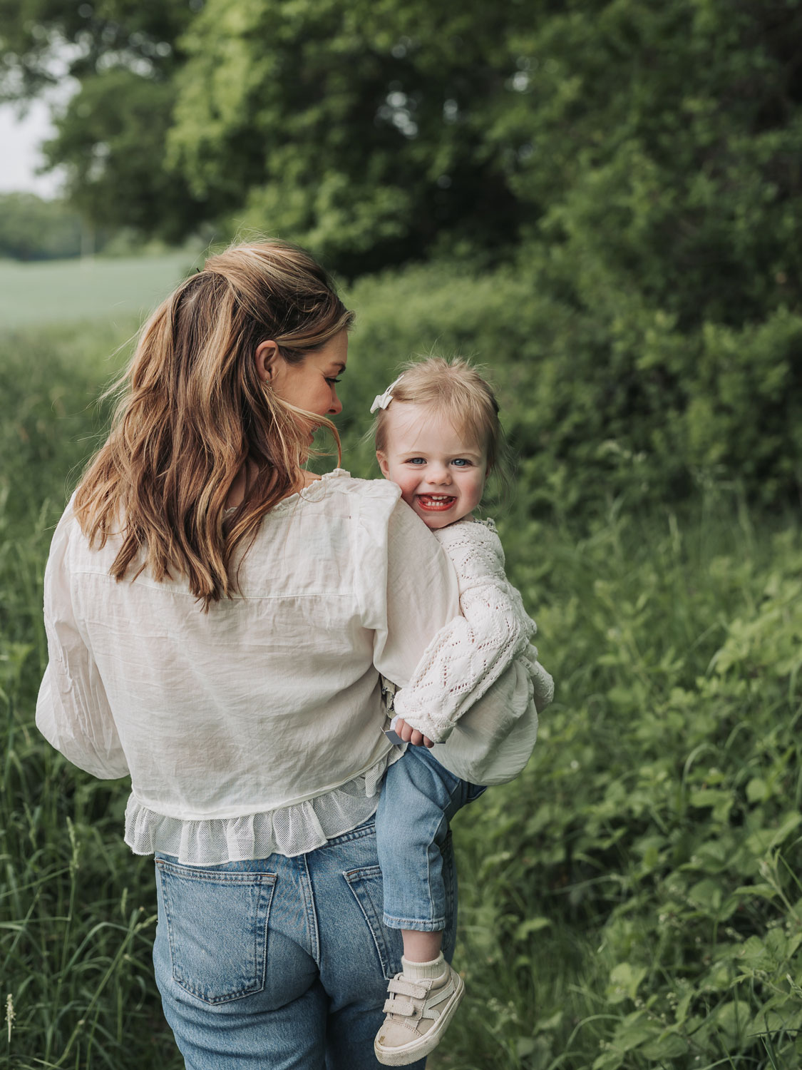 Little girl laughing at camera over mum's shoulder during a crop field photoshoot in Warwickshire