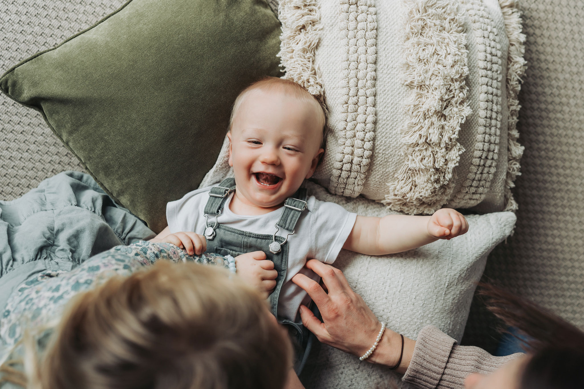 Toddler boy lying on cushions and laughing being tickled at in home newborn photoshoot by The Corbett Creative Photography