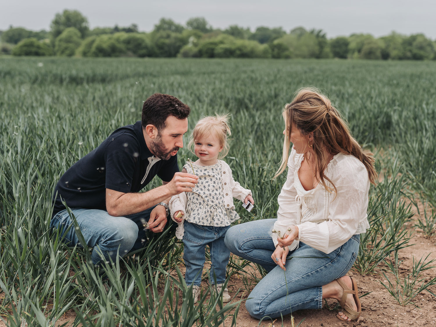Family in crop field blowing dandelions with little girl by The Corbett Creative Photography