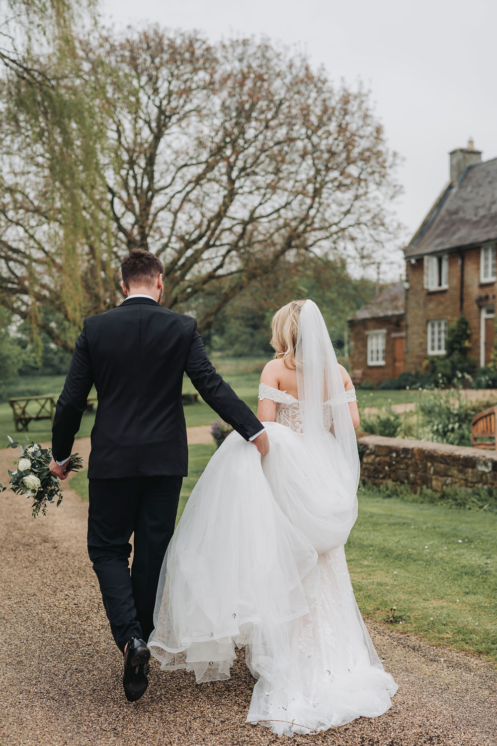 Wedding couple walking away from camera with groom holding brides dress up at Crockwell Farm wedding venue by The Corbett Creative Photography