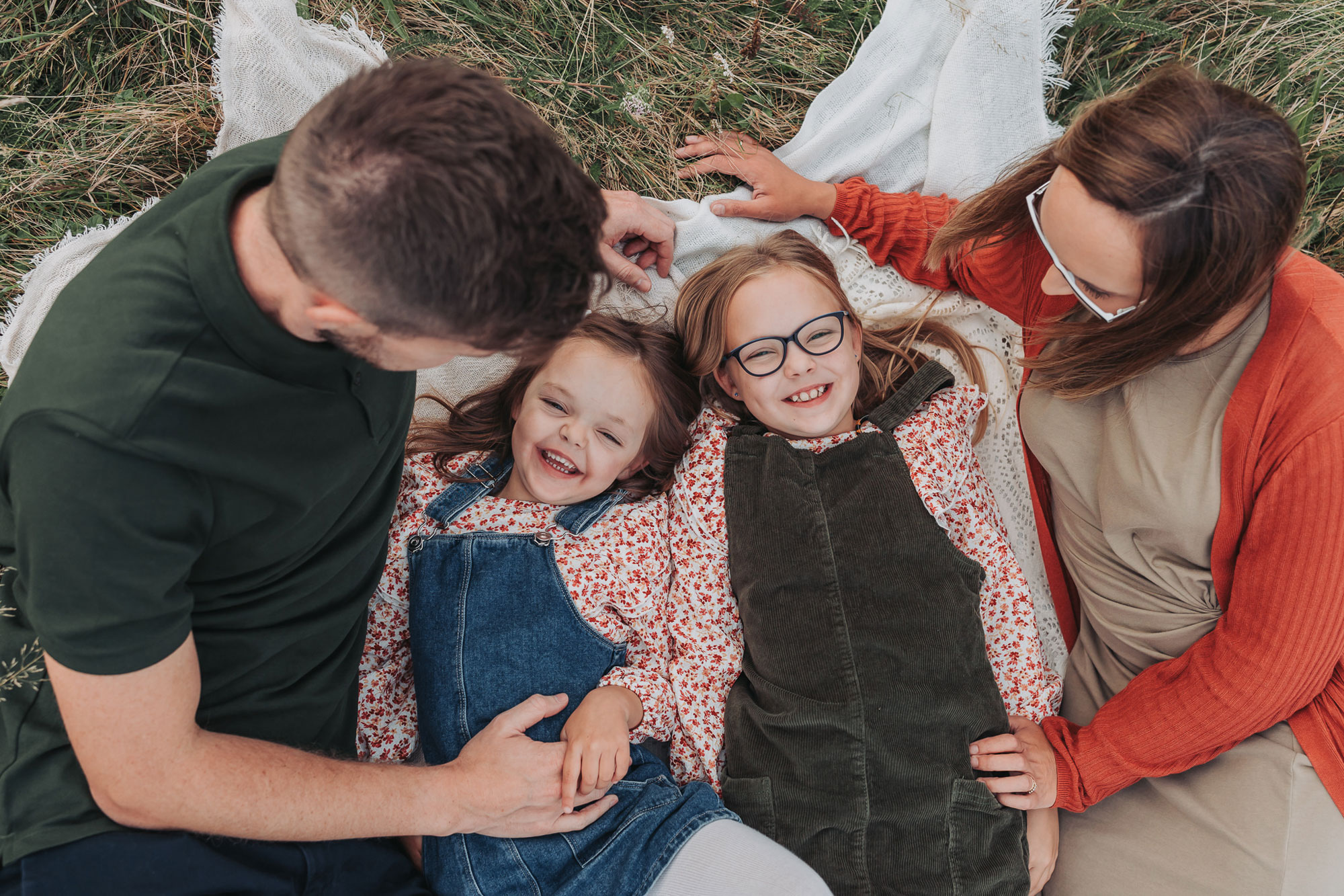 Girls wearing floral outfits and dungaree dresses lying down and smiling at the camera with parents each side during an outdoor photoshoot in Warwickshire