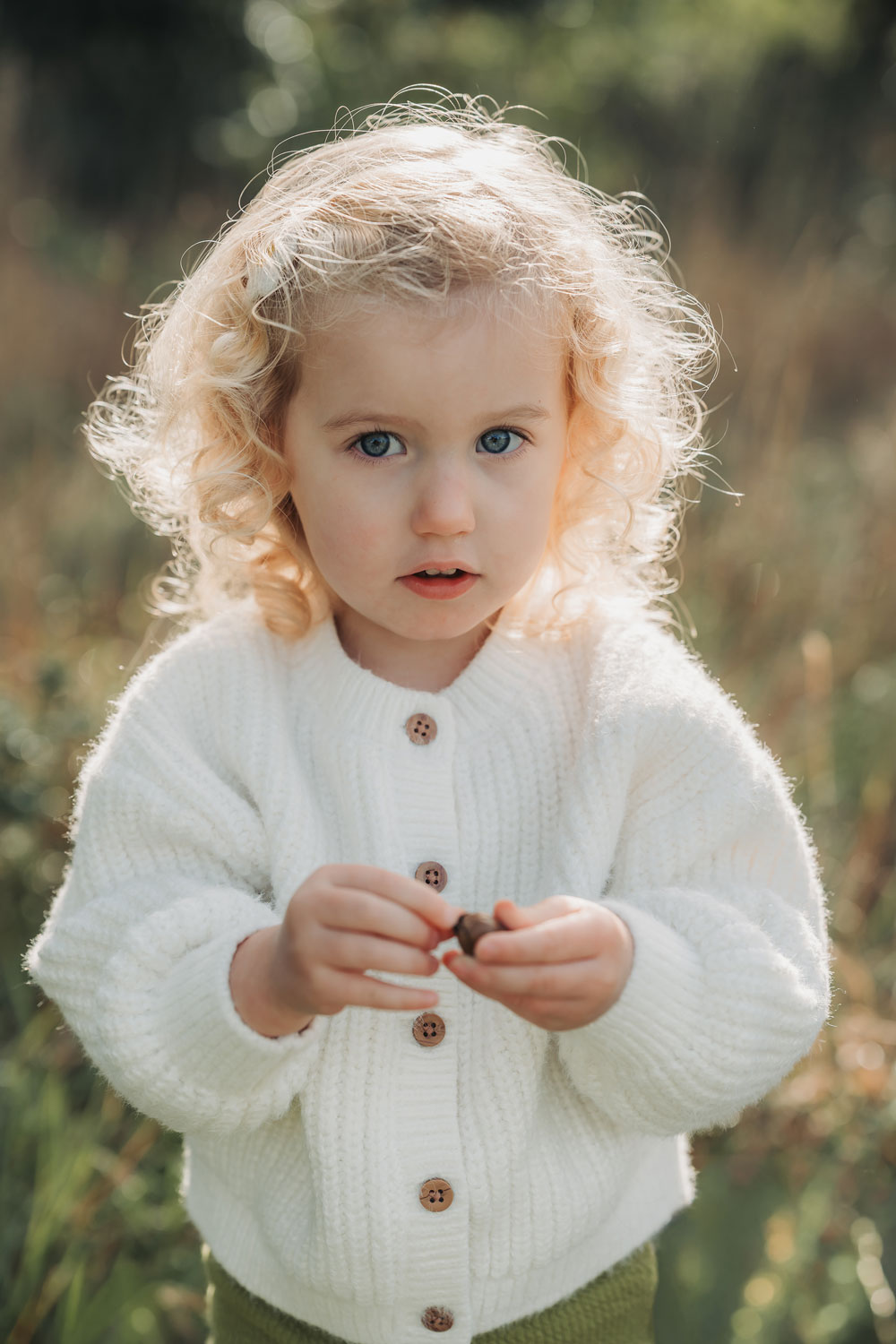 Little girl with blonde girl hair and wearing a white knitted cardigan looking up at camera during an outdoor family photoshoot in Stoneleigh, Warwickshire