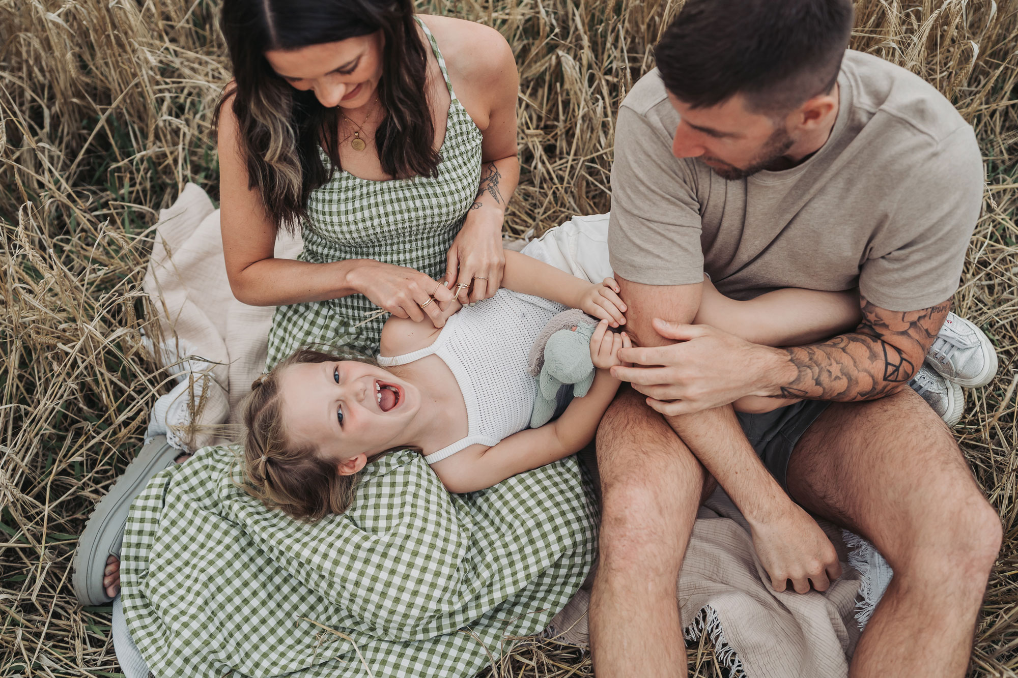 Crop field golden hour family photoshoot with little girl lying down being tickled
