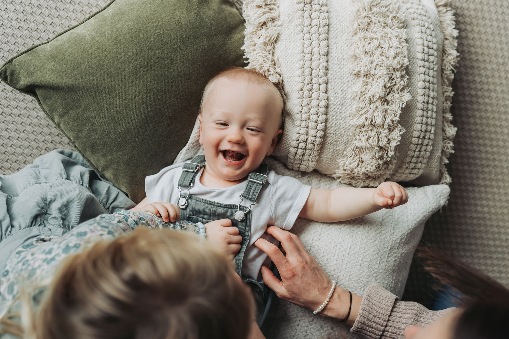 In home family photoshoot with baby boy lying on cushions and smiling up at camera
