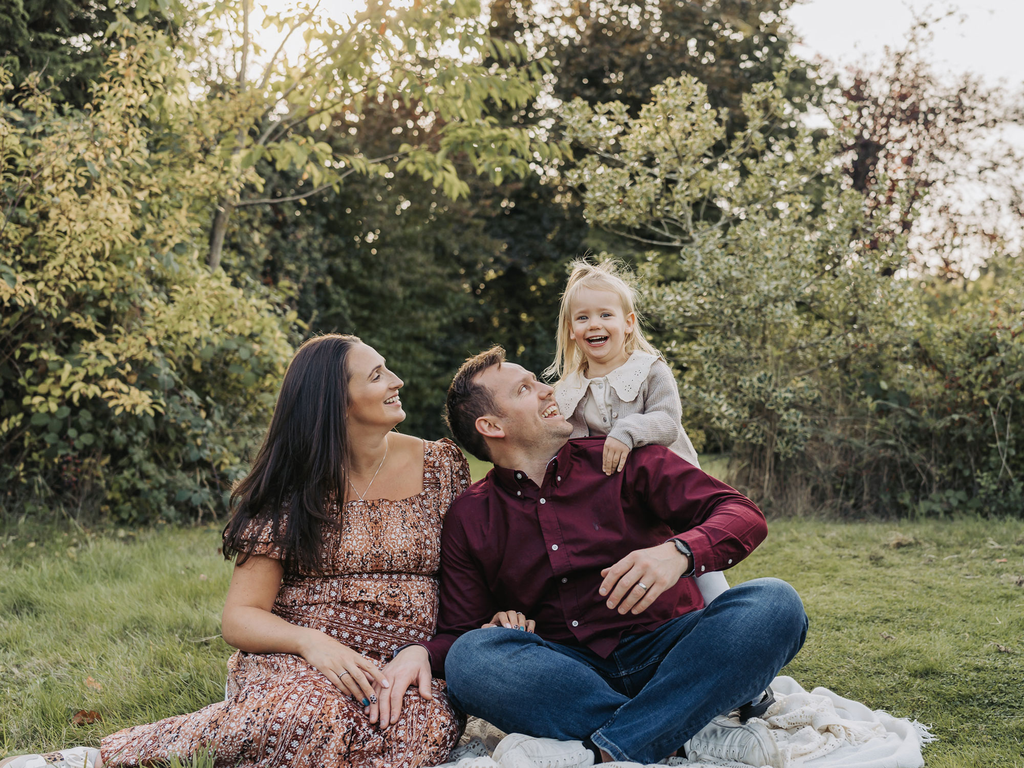 Outdoor golden hour maternity photoshoot with little girl popping up behind parents and smiling