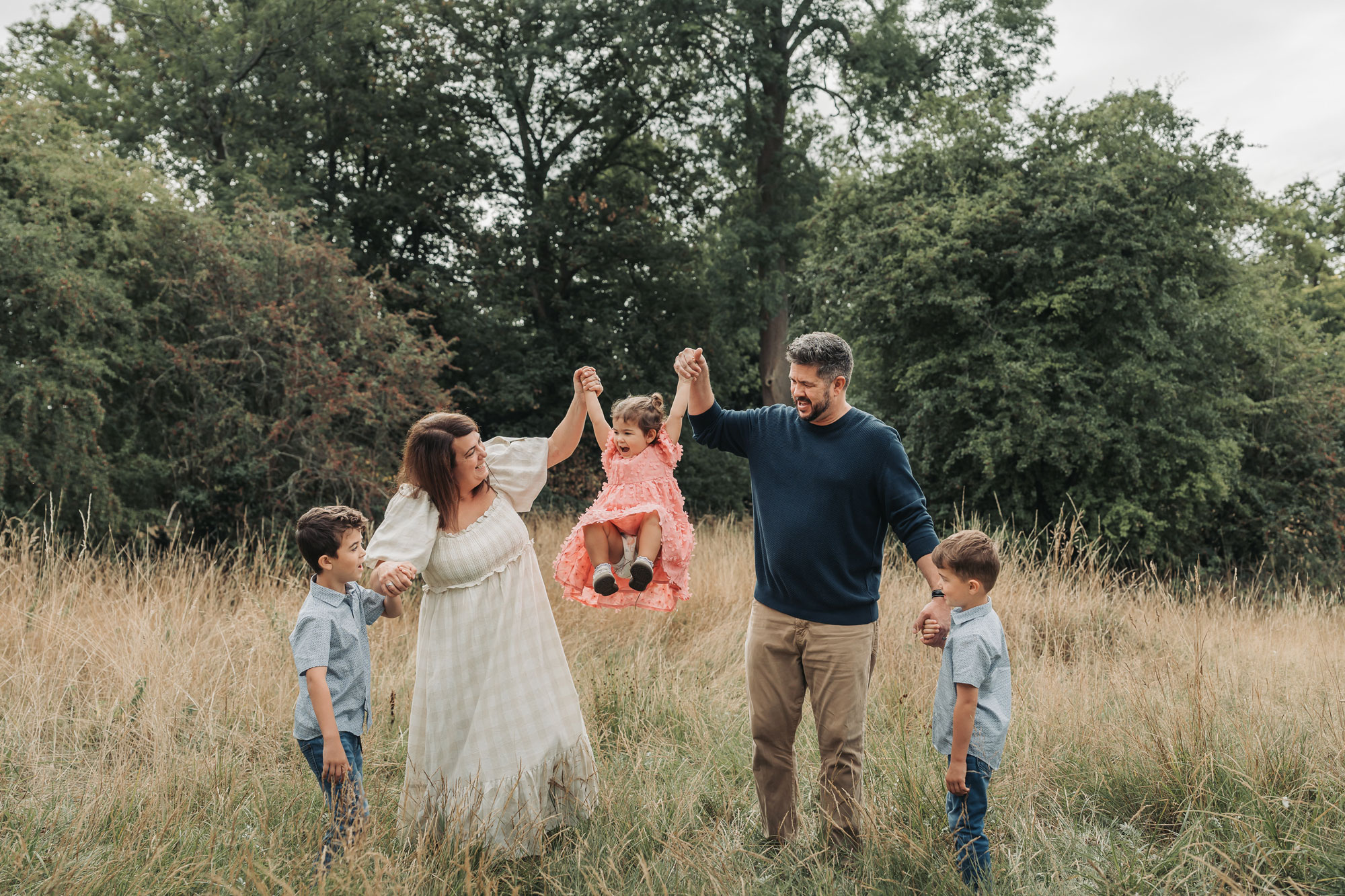 Family of five in Kenilworth holding hands together lifting toddler daughter up in long grass
