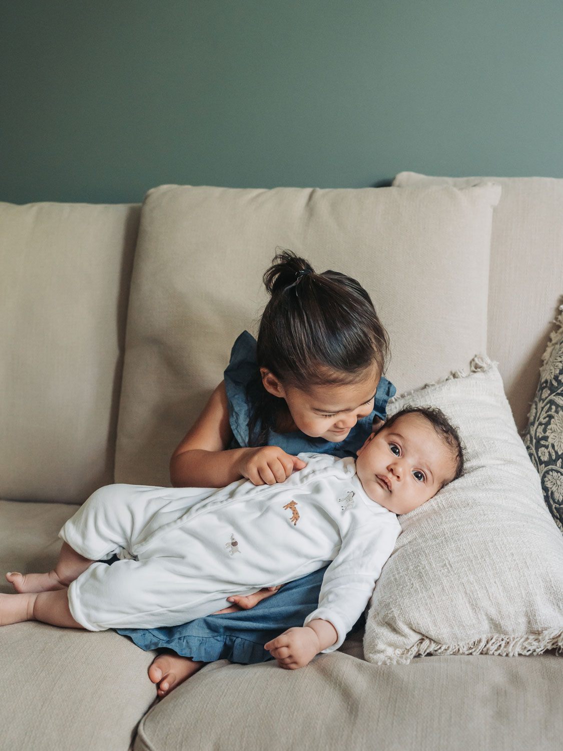 Toddler girl wearing blue dress sat on sofa holding baby sister in animal babygrow