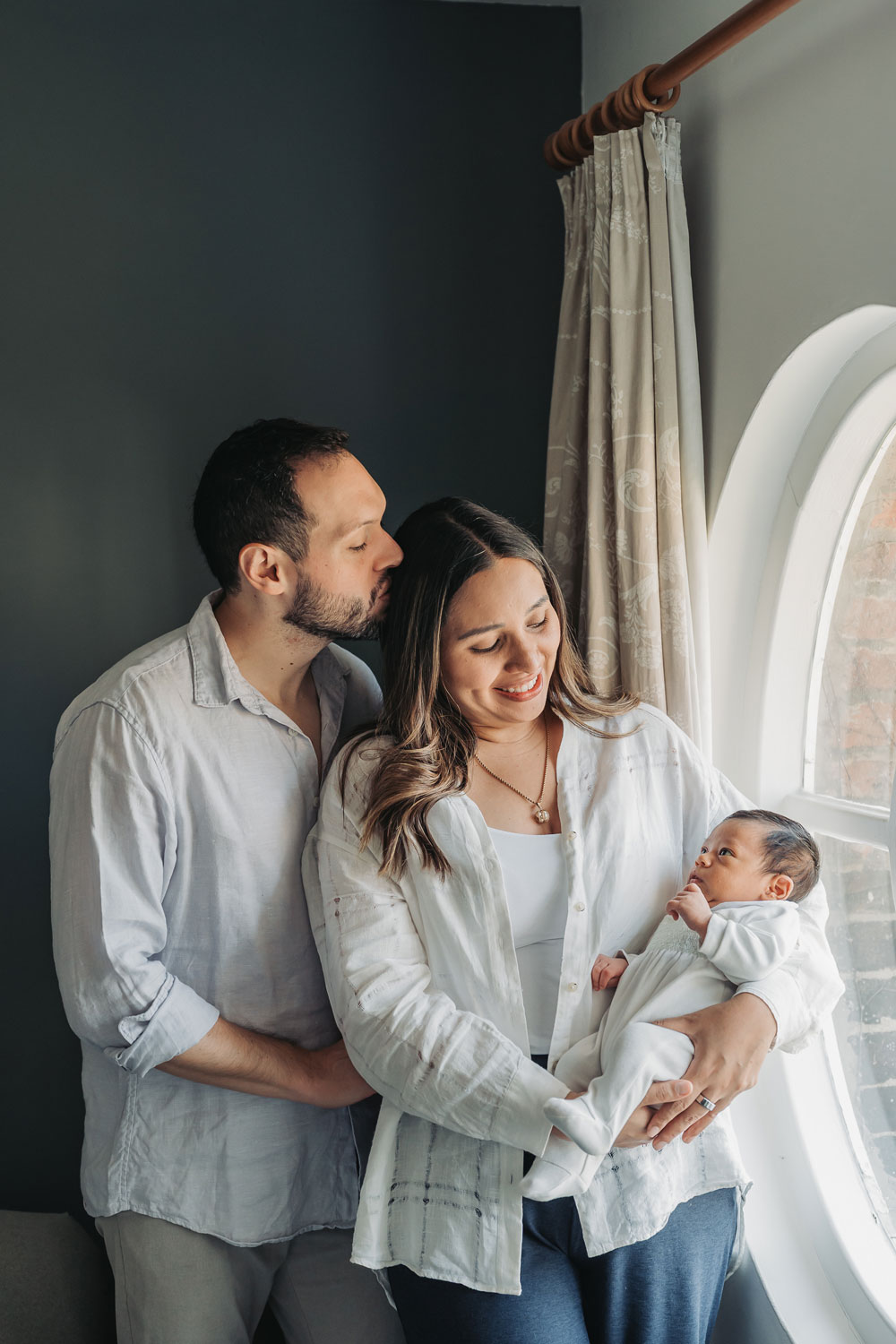 Family stood in window holding baby whilst dad kisses mum on the head