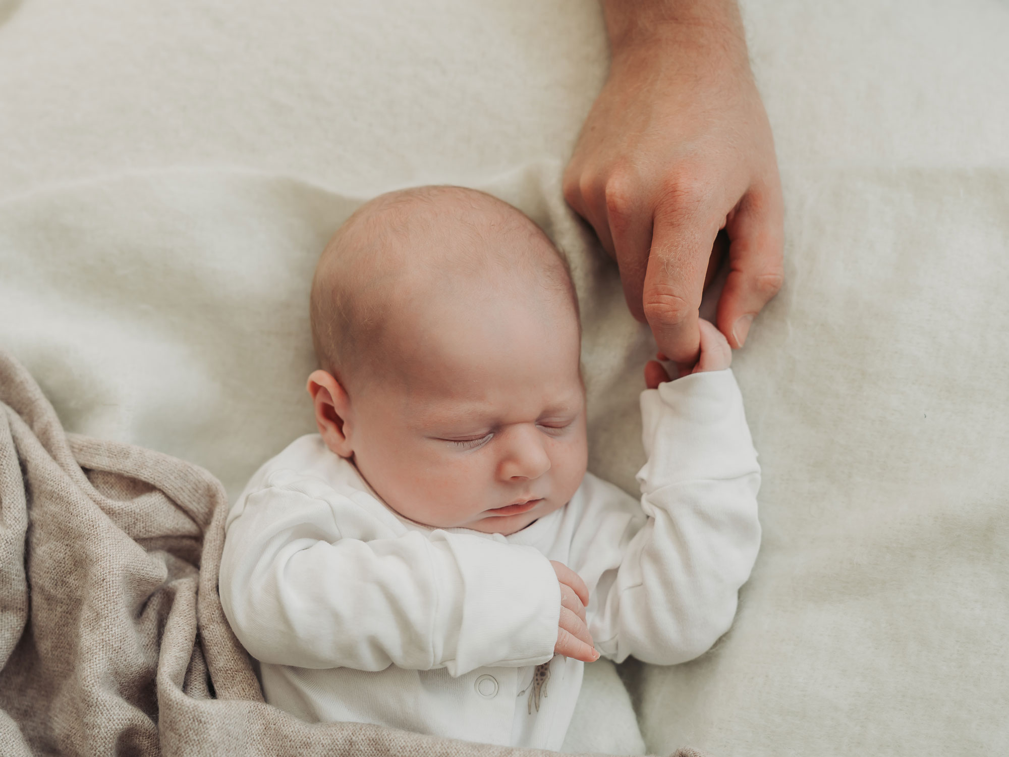 Baby wearing white babygrow asleep holding parents finger