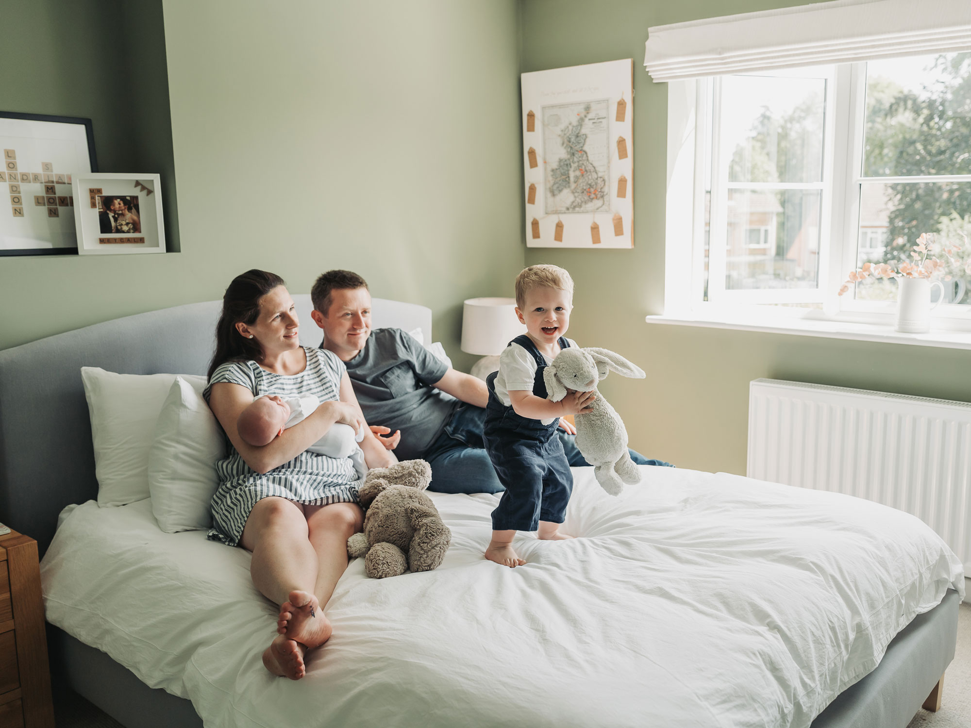 toddler in navy dungarees jumping on bed with bunny whilst parents look on