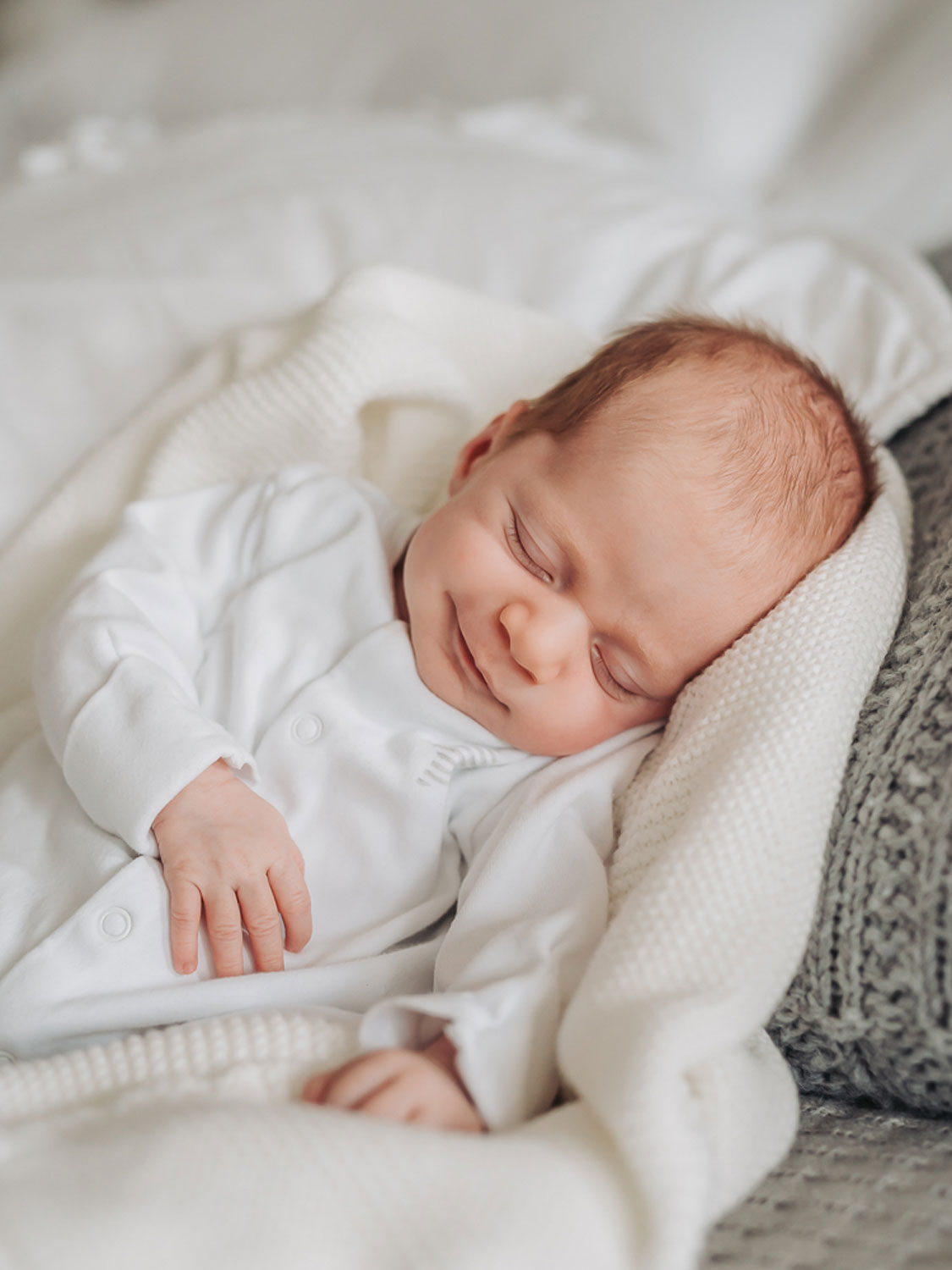 baby lying on white blanket wearing white babygrow smiling