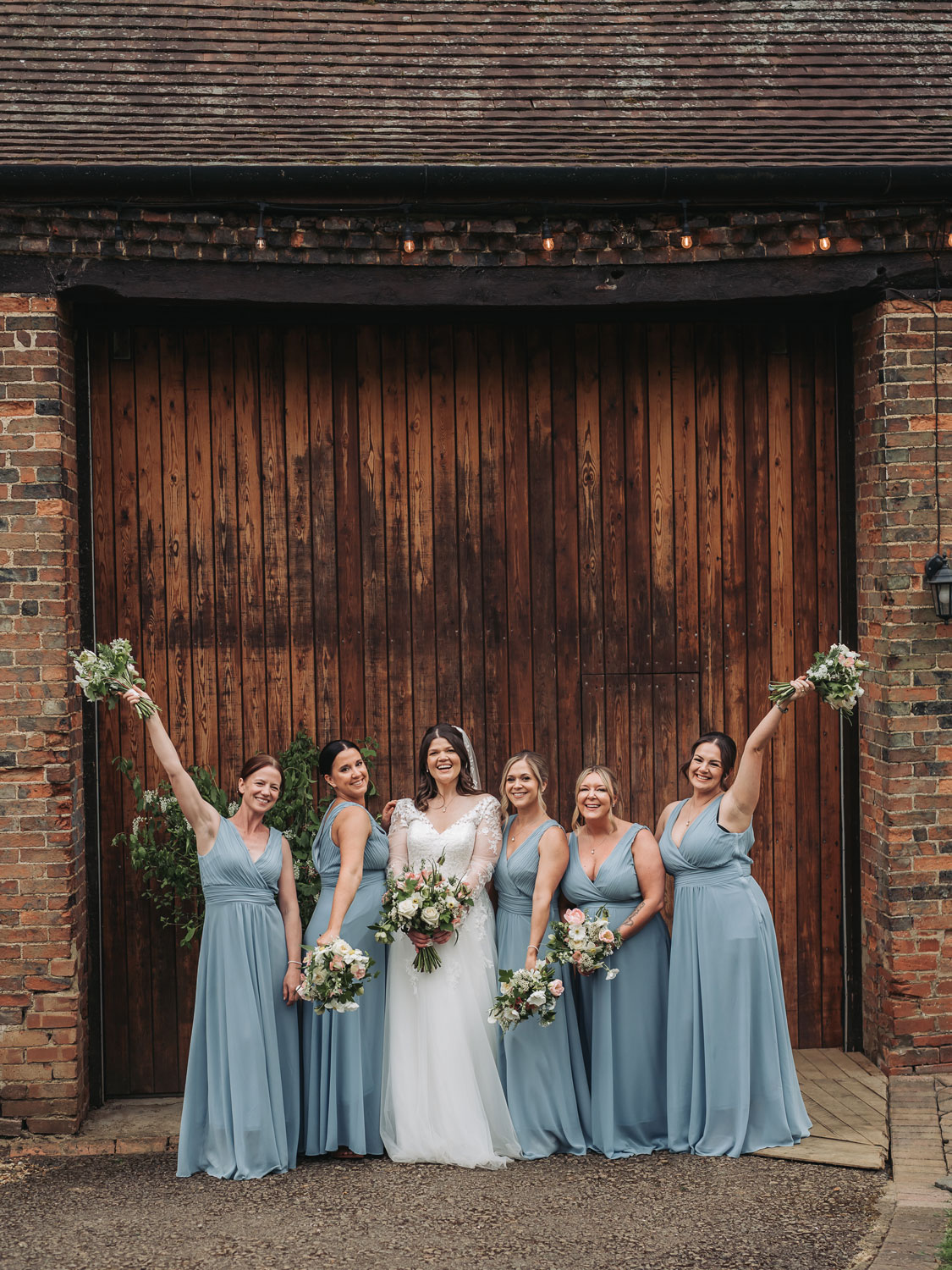 Bridal party group shot outside wooden barn doors with wedding bouqets in the air