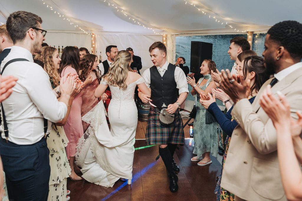 Bride and groom dancing arm in arm whilst guests clap