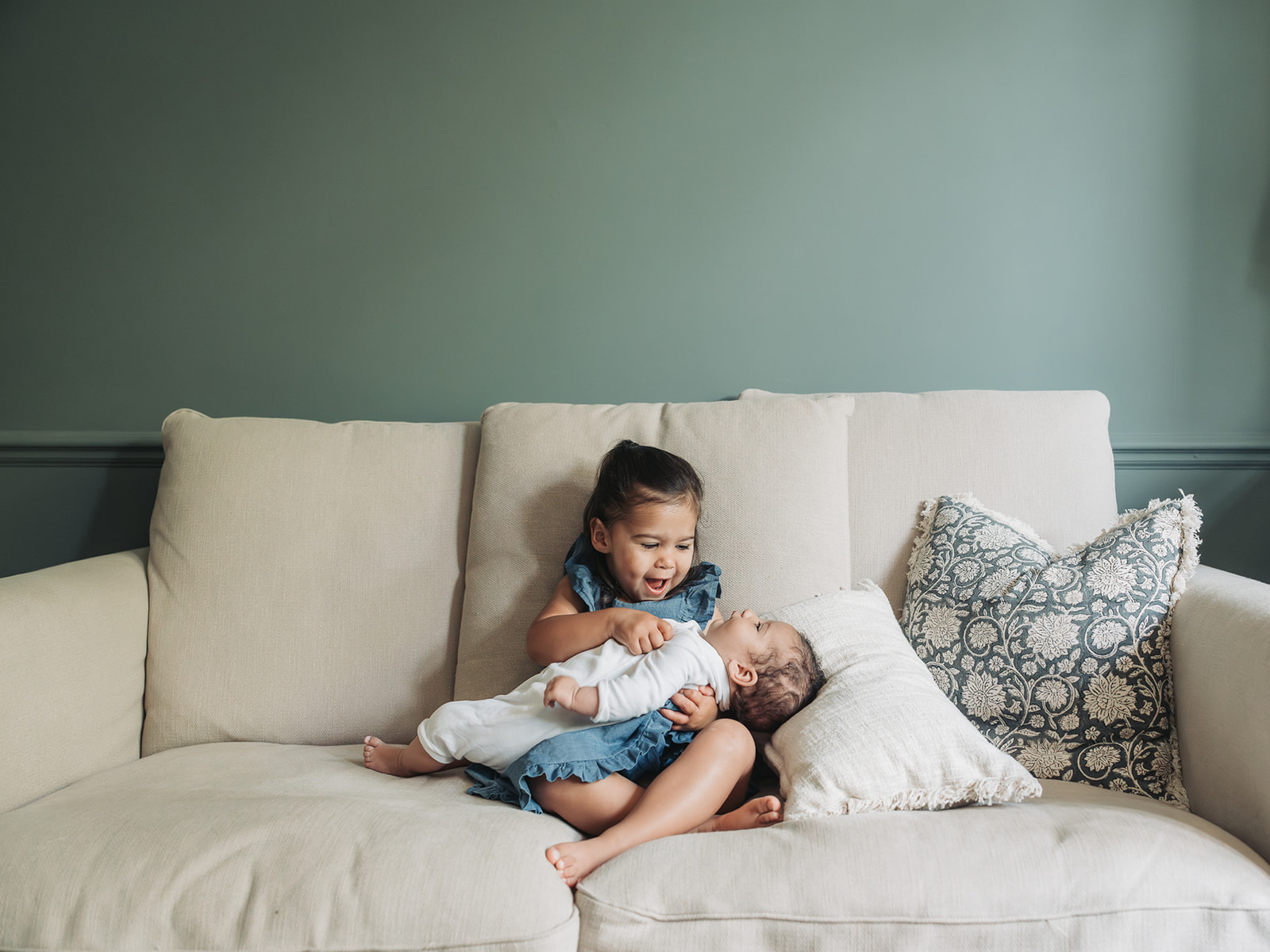 Toddler girl sat on white sofa holding baby sister and looking happy