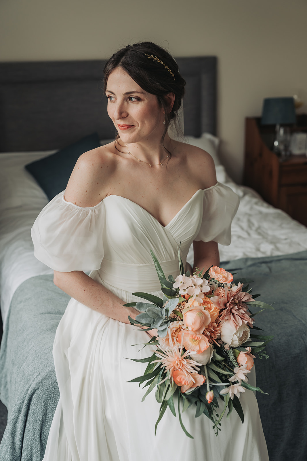 Bride sat on bed gazing to the side wearing off the shoulder wedding dress holding peach bouquet