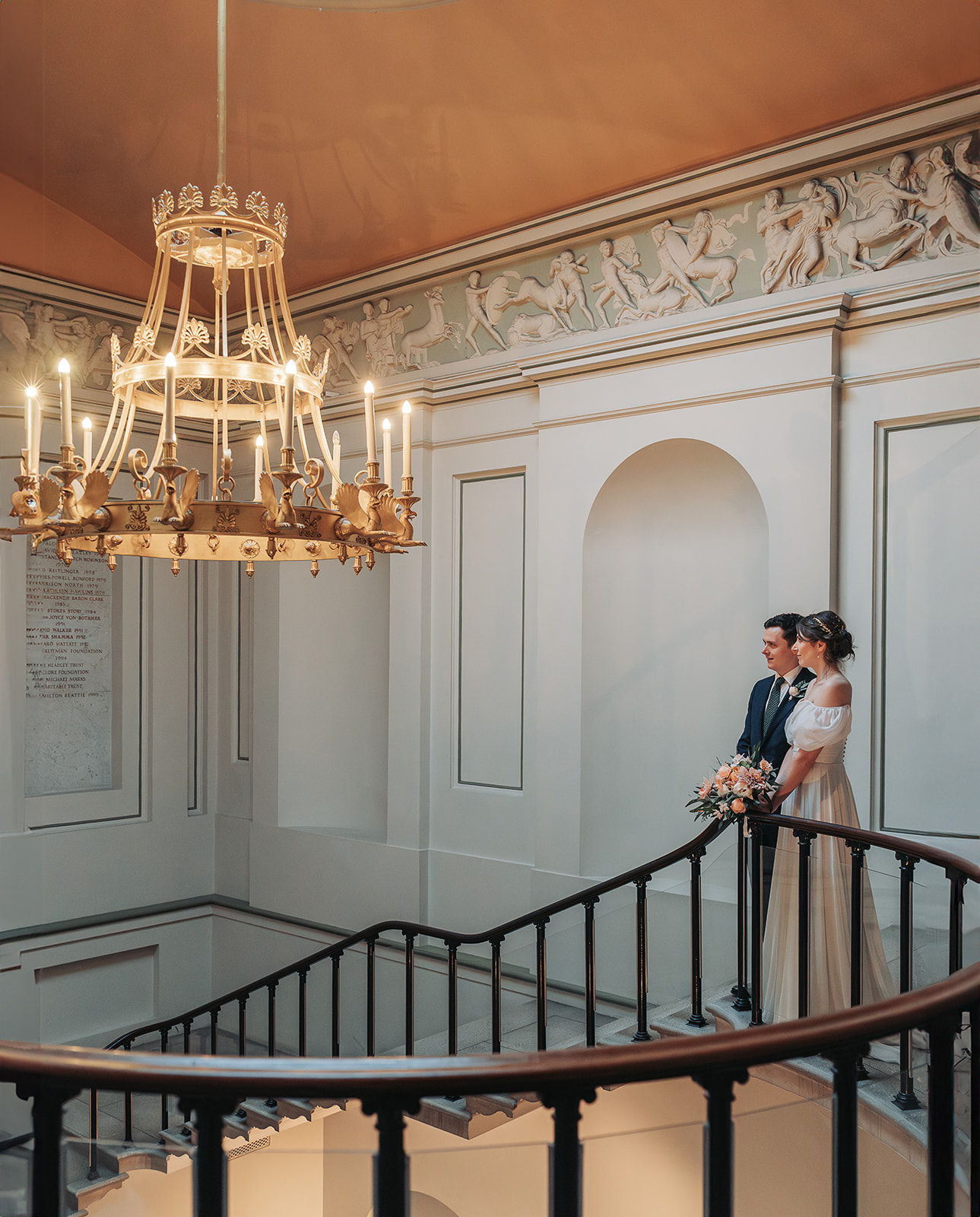 Bride and groom stood at top of grand staircase with chandelier