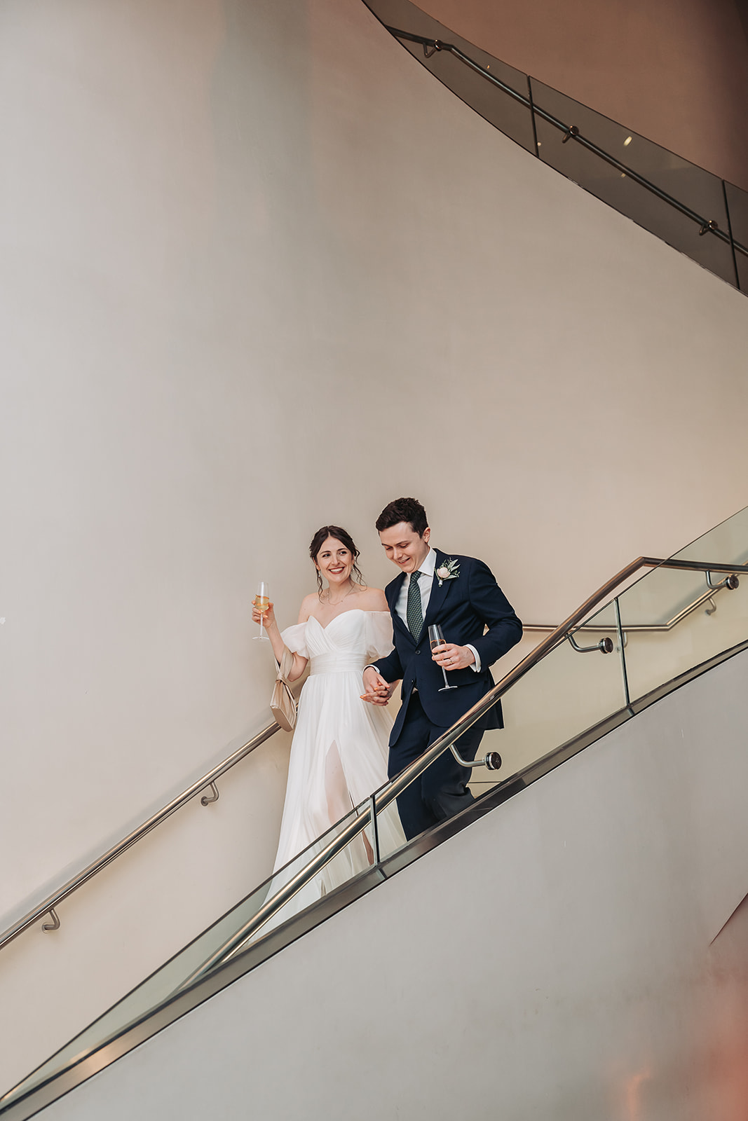 Bride and groom walking down atrium stairs holding champagne glasses