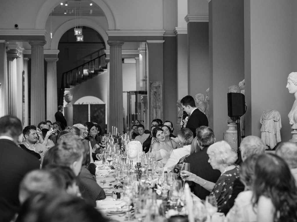 Black and white photo of bride looking at groom giving speech
