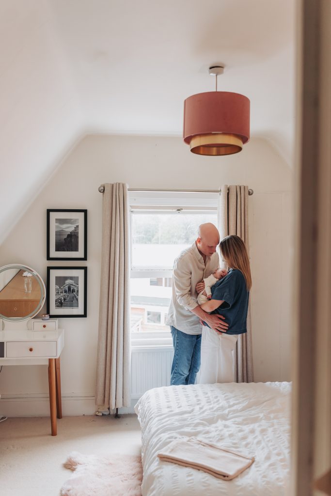 Mum and dad cuddled by window with newborn baby 