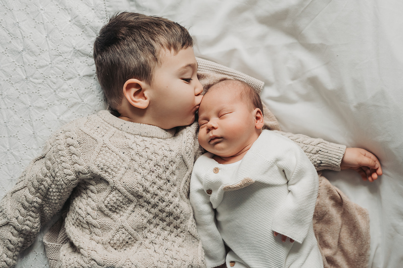 Toddler boy in knitted beige jumper kissing sleeping baby in newborn photoshoot