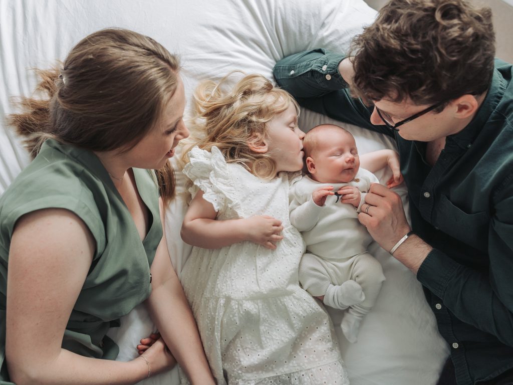 Family lying on bed with toddler girl kissing newborn baby
