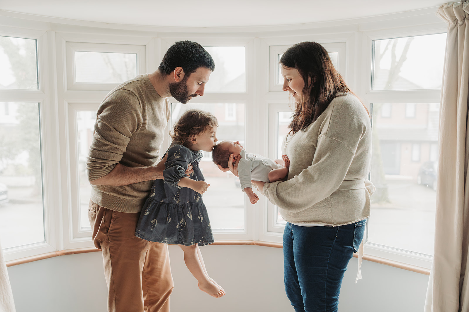 Family in front of window with toddler girl being lifted up to kiss new baby brother on head
