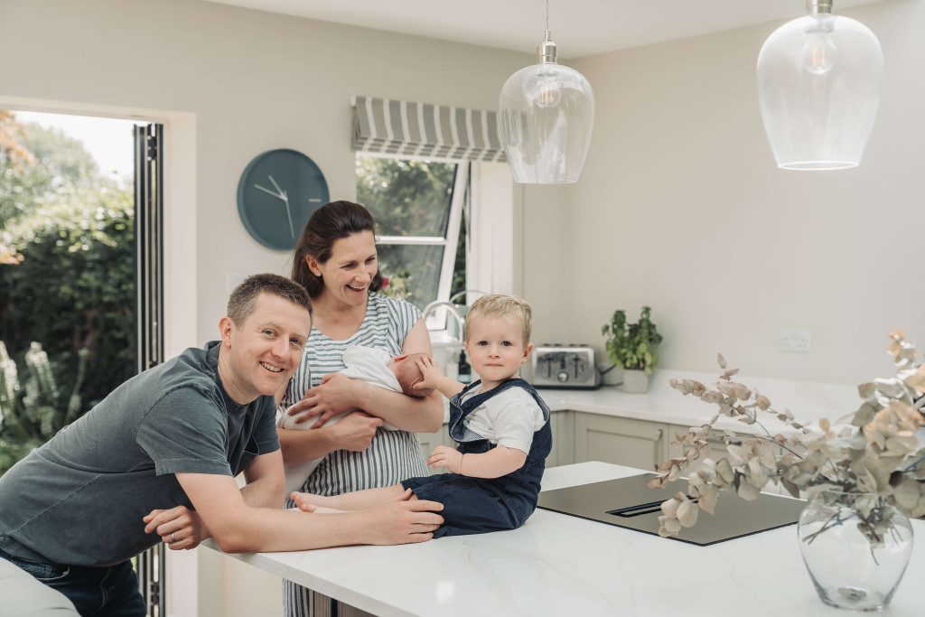 Toddler boy stroking baby's head whilst sat in the kitchen at home in Kenilworth