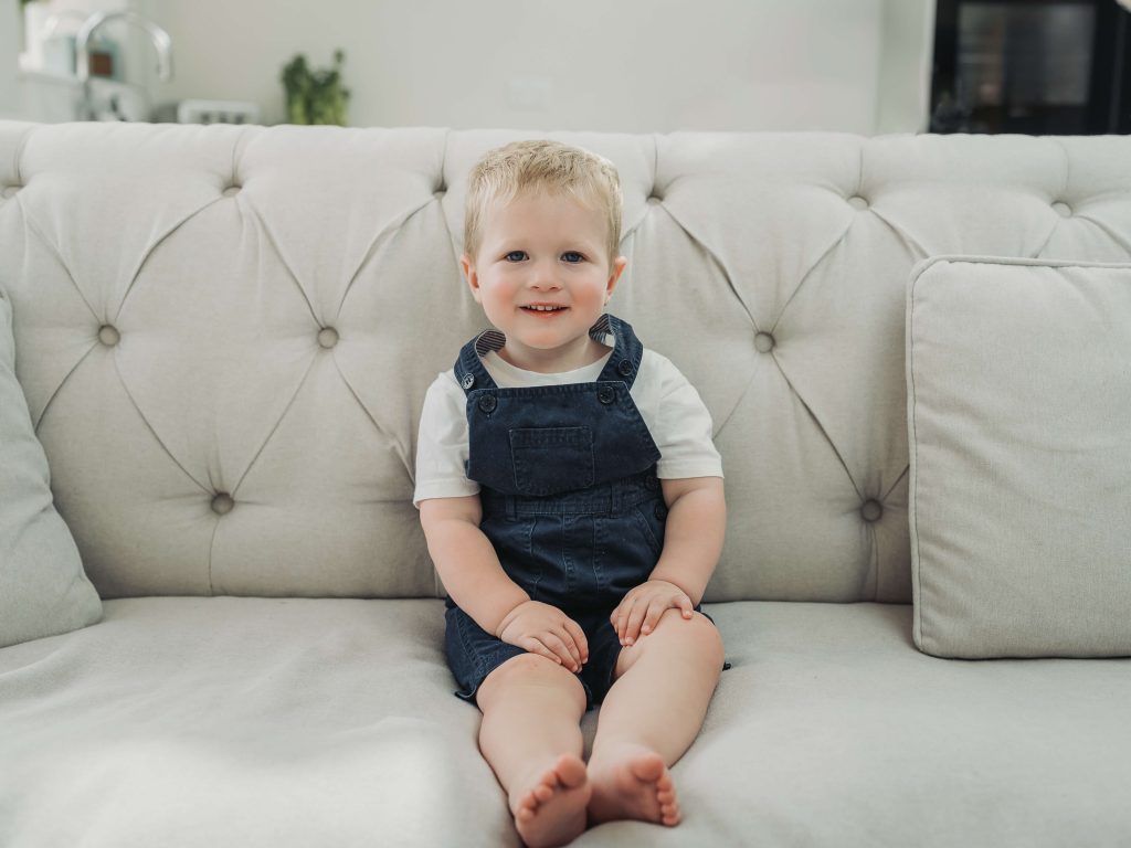 Toddler boy in navy dungarees sat on sofa smiling at camera during an in home photoshoot