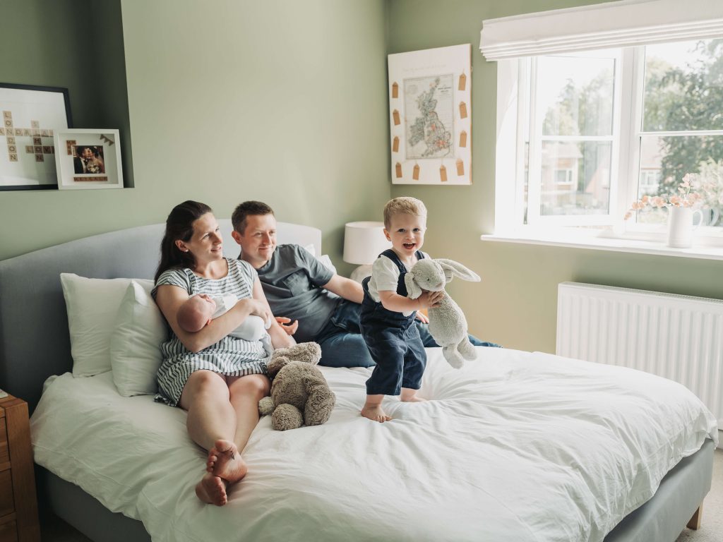 Toddler boy holding fluffy bunny and jumping on the bed whilst family watch on