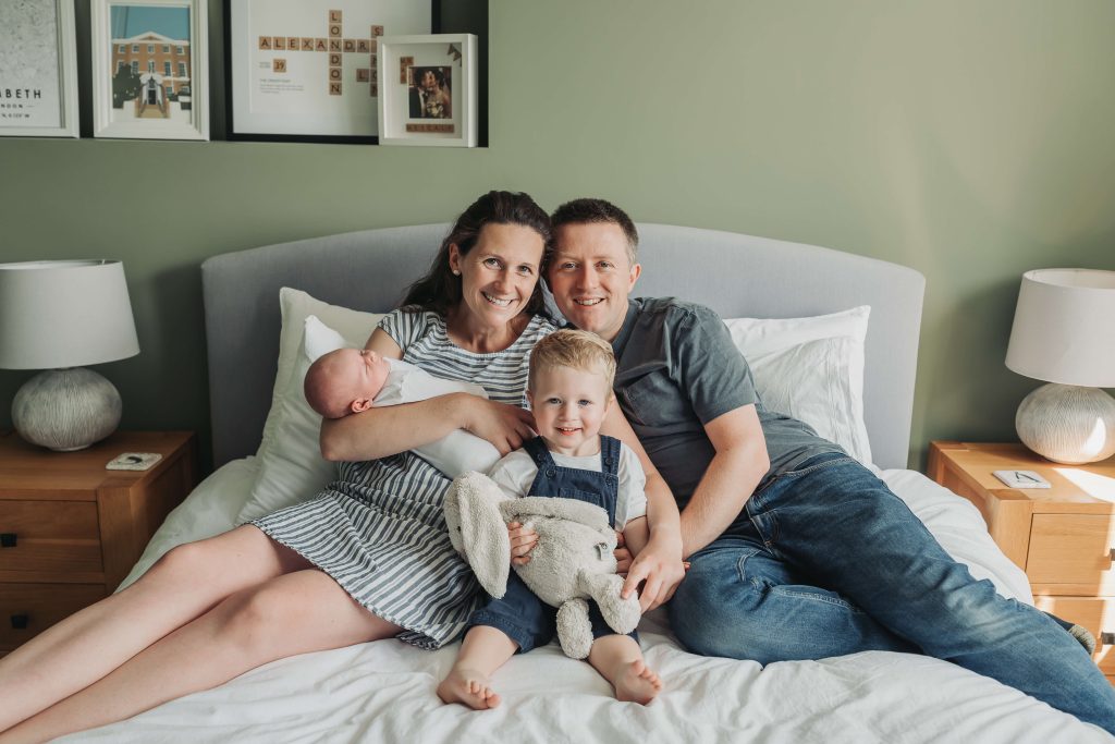 Family with toddler and baby smiling at the camera on the bed during an in home newborn photoshoot in Kenilworth