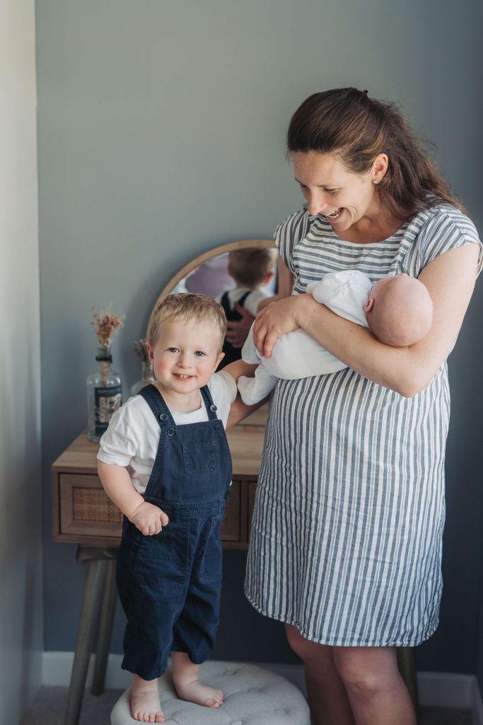 toddler boy smiling at the camera whilst mum holds baby during in home Kenilworth photoshoot