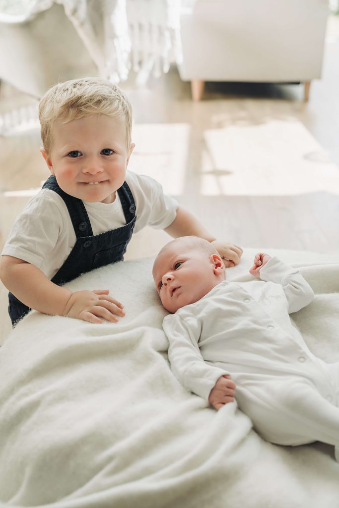 toddler boy looking at camera whilst kneeling down next to baby brother