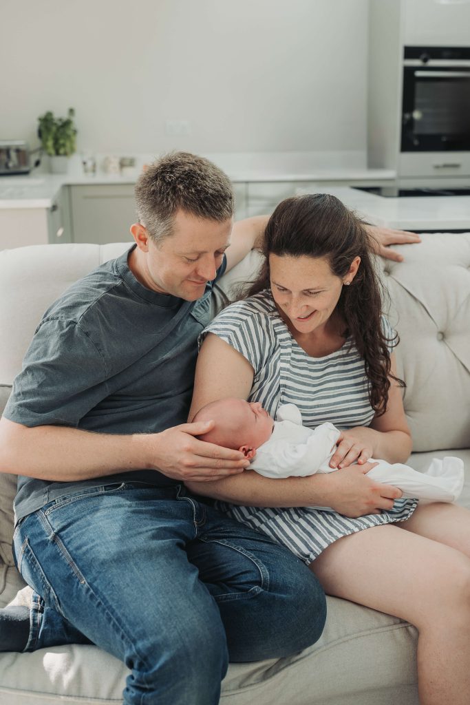 Mum and dad looking down at baby in white babygrow during newborn photoshoot