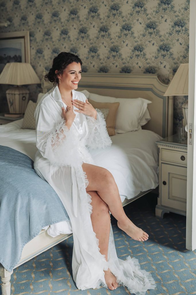 bride in white dressing gown sat on bed smiling
