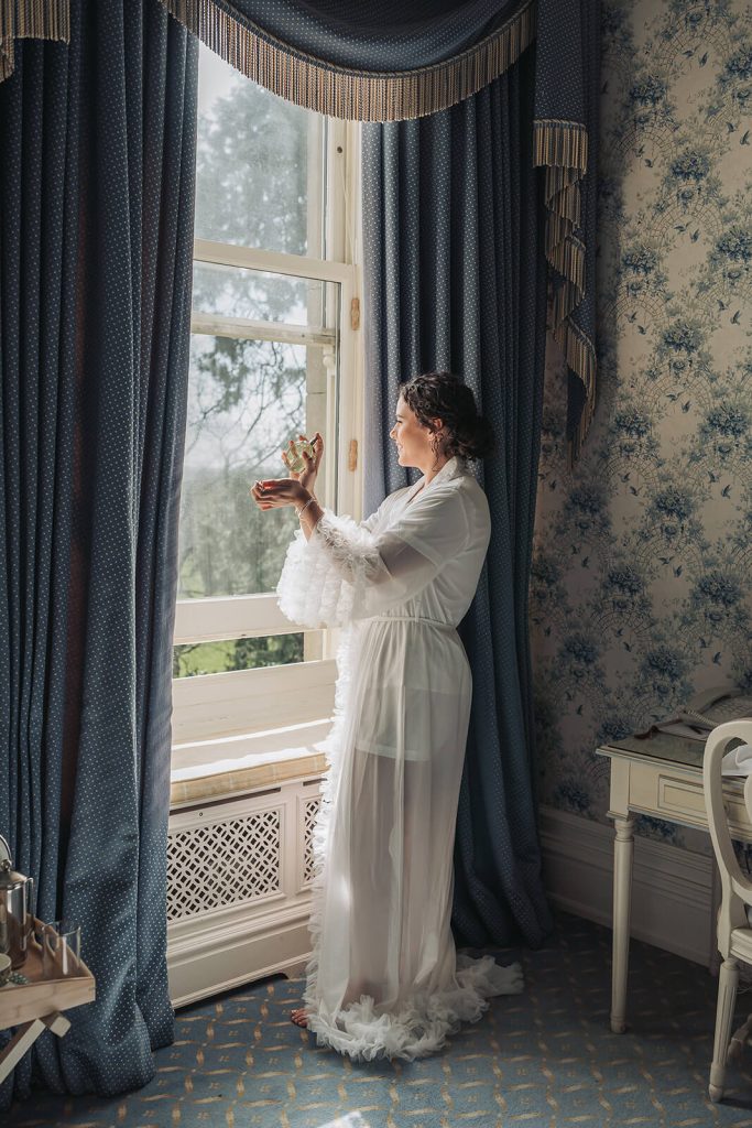 bride in dressing gown looking out of the window putting on perfume