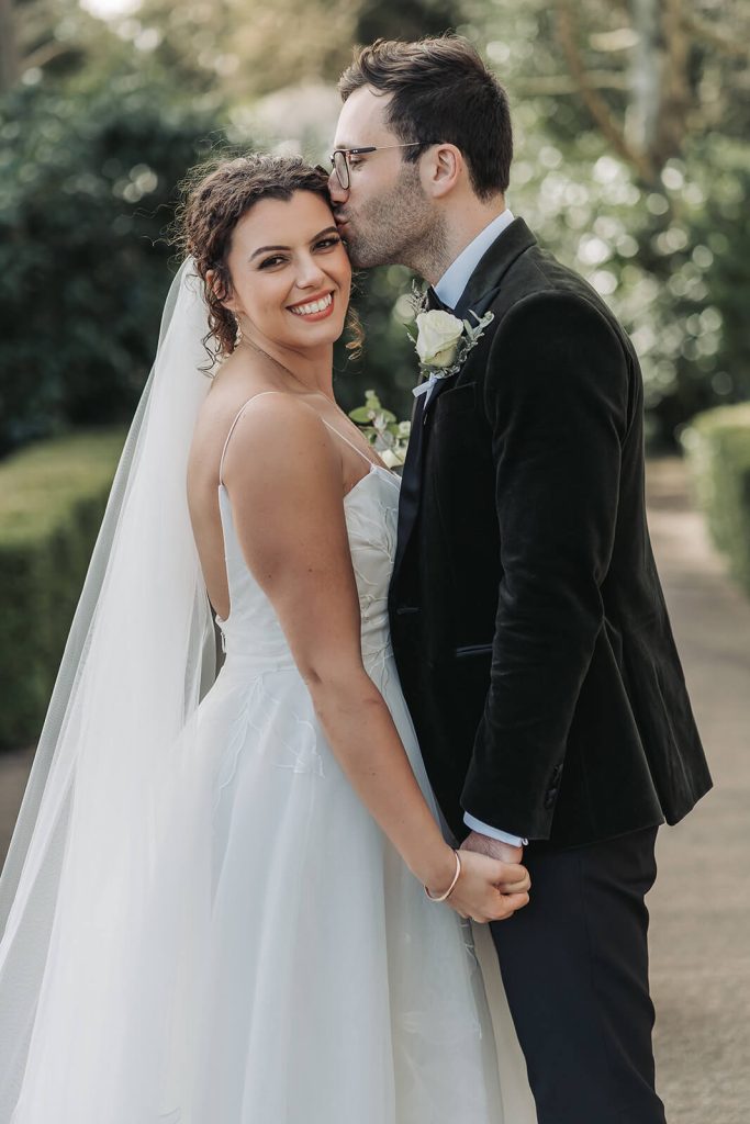 bride looking at camera whilst groom kisses the side of her head