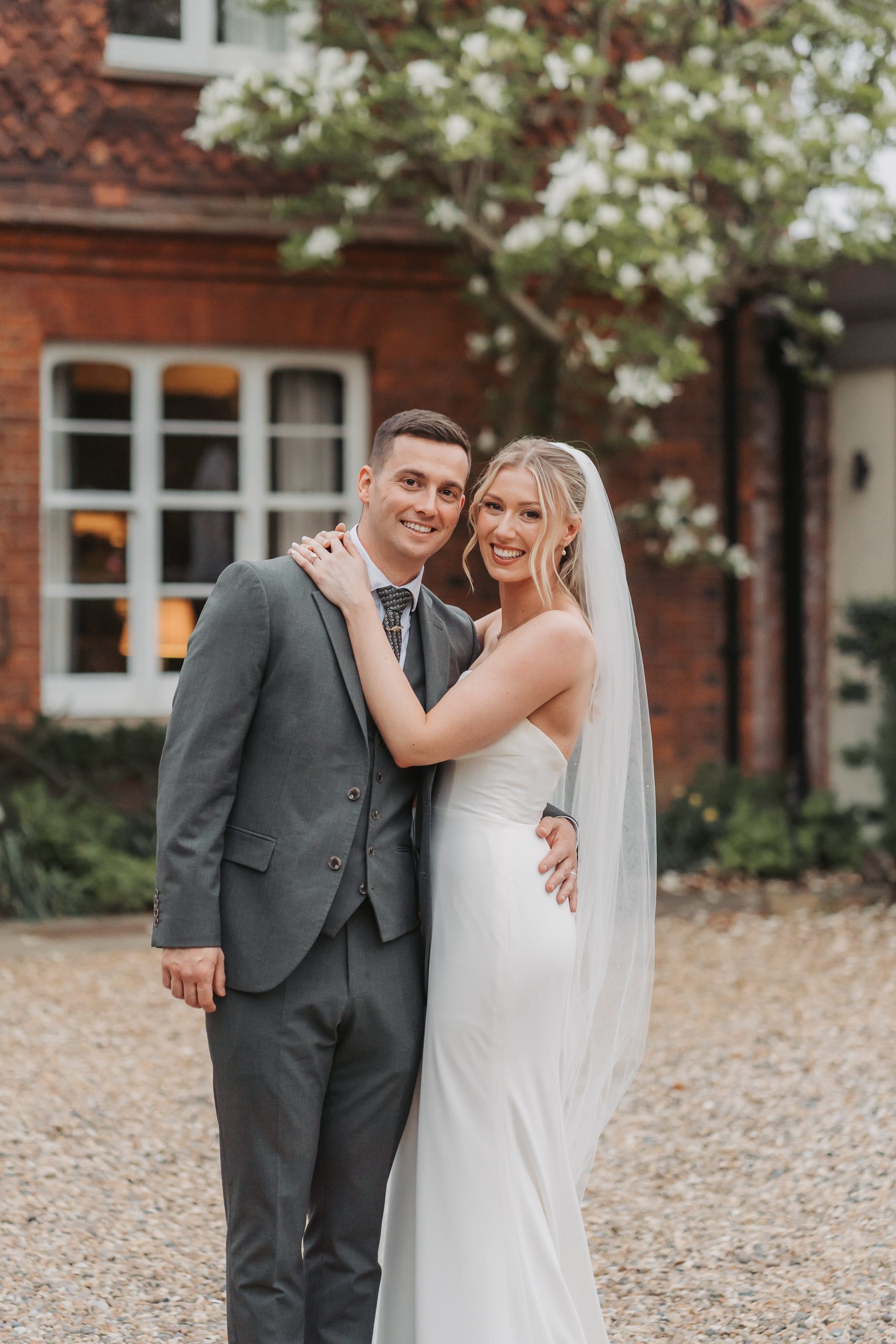 Bride hugging groom and smiling at the camera with blossom tree behind at Farmhouse at Redcoats